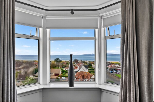 A bay window with a clear view of a coastal landscape, including houses and the sea. - Charming Devon Cottage With Garden (West Kilbride)