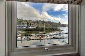 Vista a través de una ventana de un puerto deportivo con barcos amarrados, cielo despejado y un paisaje montañoso al fondo.