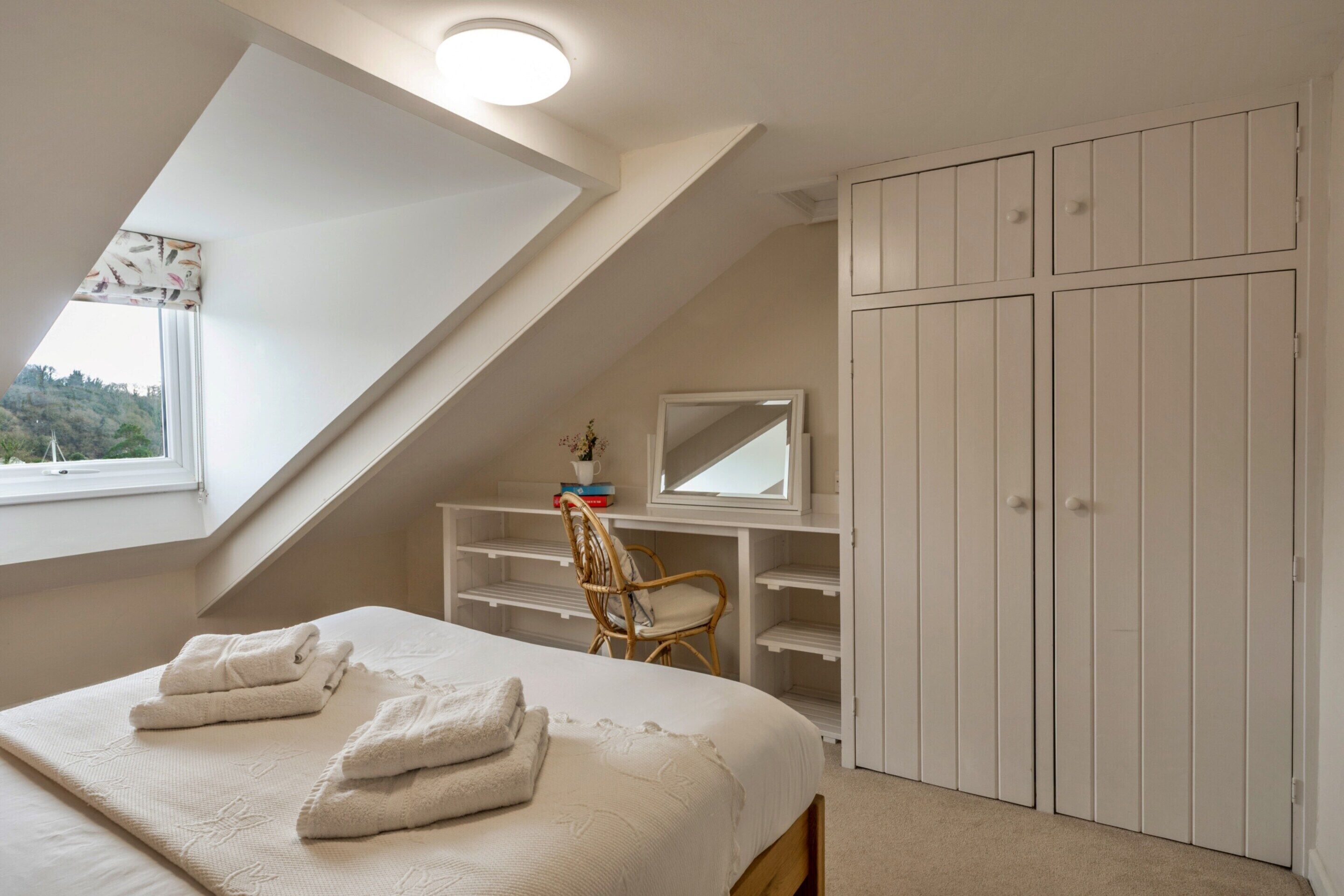 A bedroom with a bed, a chair, a desk with a mirror, and a wardrobe.