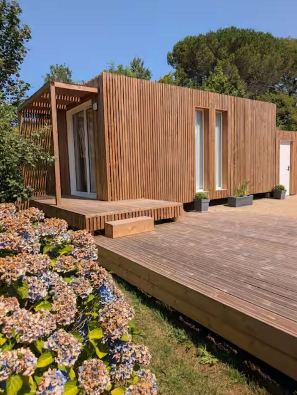 A modern wooden structure with a deck, surrounded by greenery and flowers.