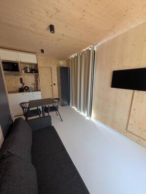 A modern living space with a sofa, dining table, chairs, a flat-screen TV, and a kitchen area with appliances. - The Landaoudec Cabin (Crozon)