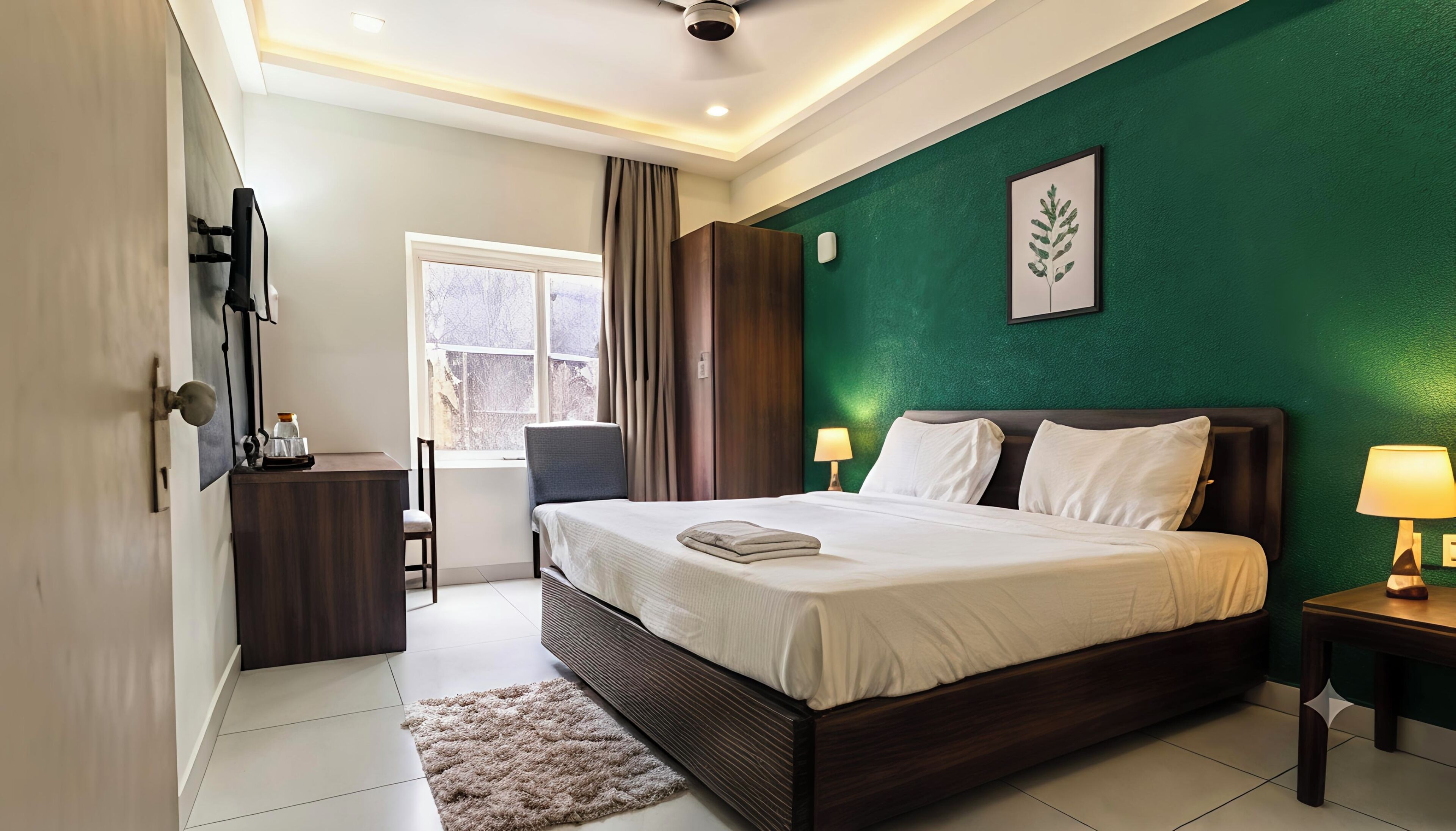 A hotel room with a bed, a desk, a chair, a window, and a green accent wall.