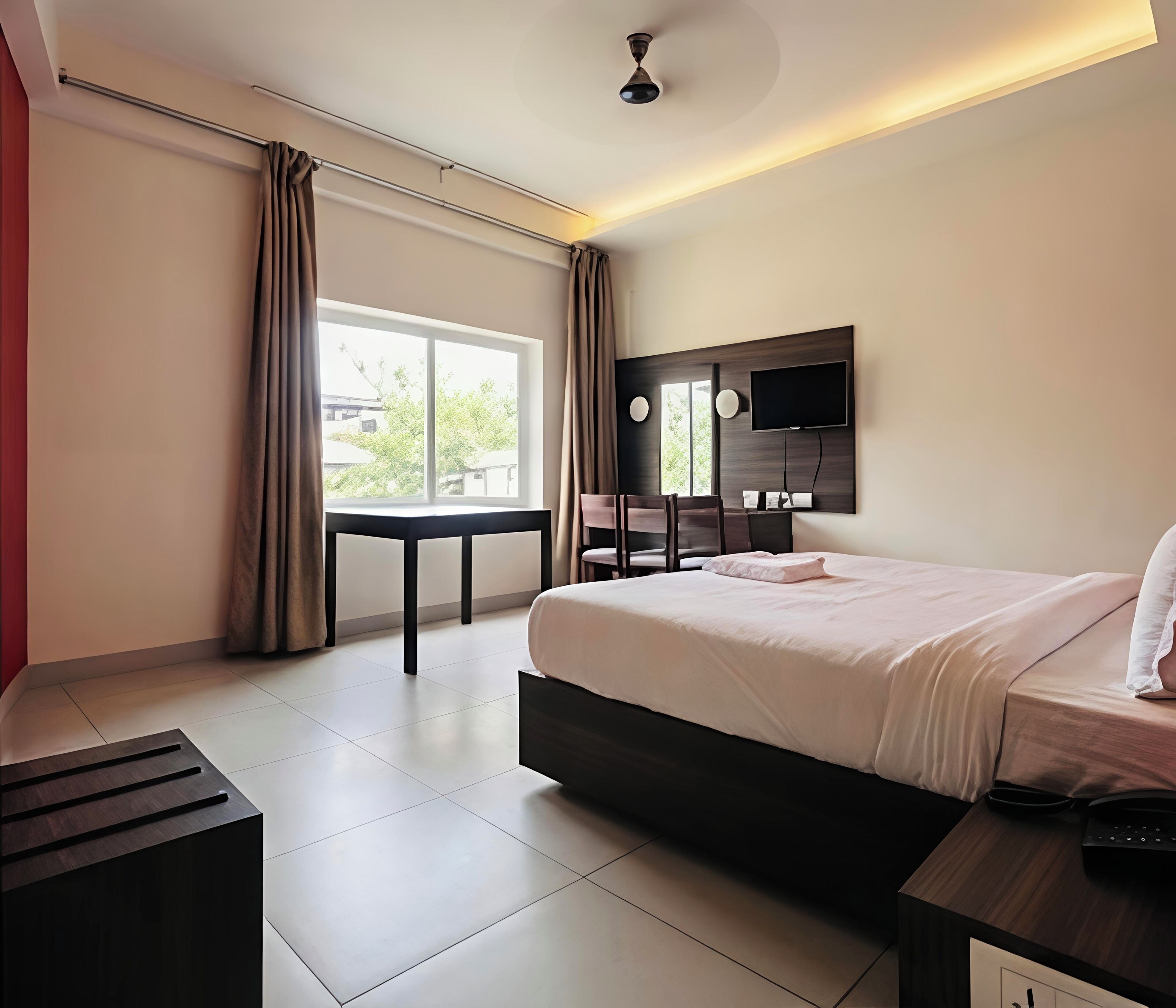 Quarto de hotel moderno com uma cama grande, uma escrivaninha, uma cadeira, uma televisão na parede e uma janela com cortinas.