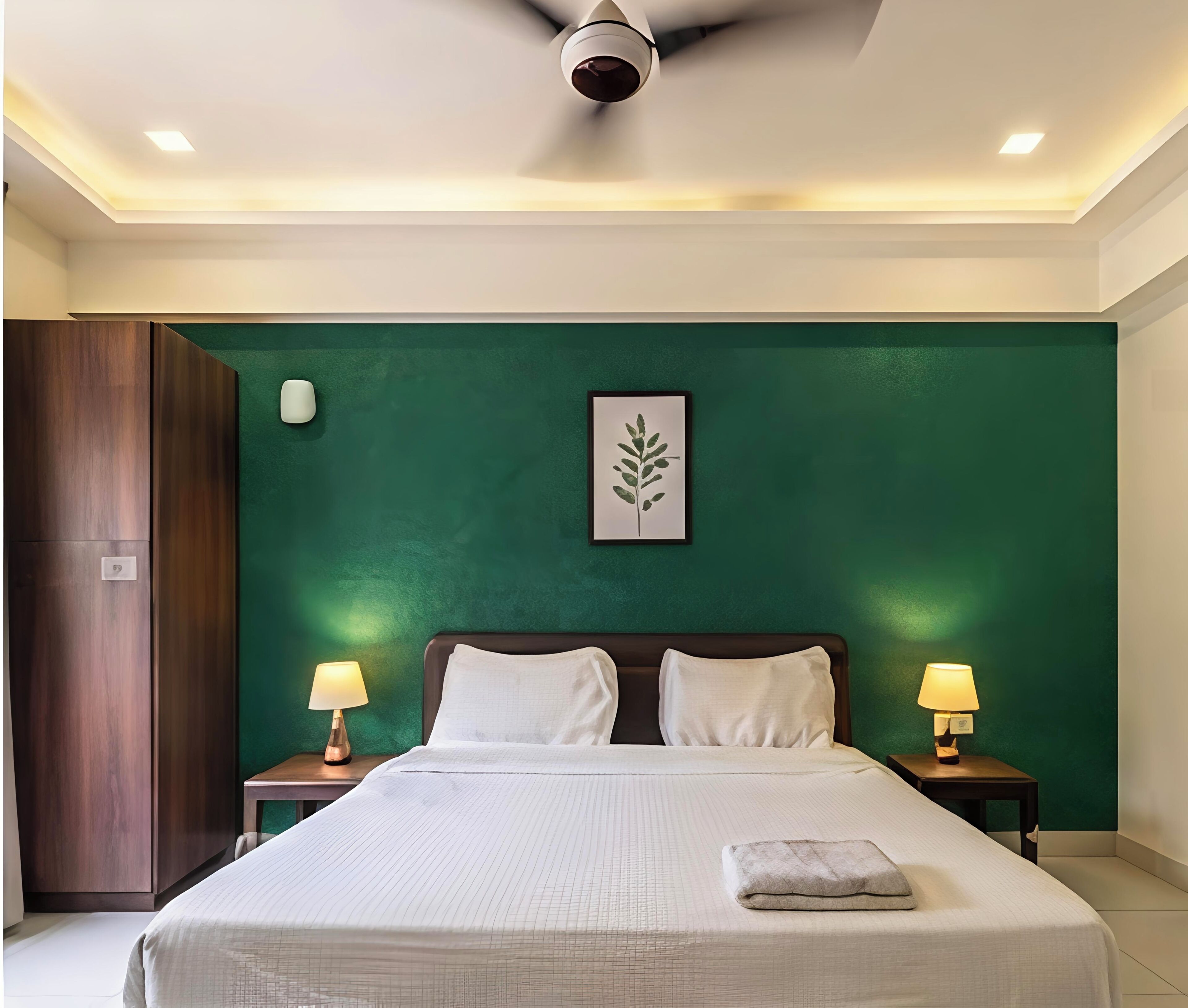 A hotel room with a bed, two bedside lamps, a ceiling fan, a green accent wall, and a framed leaf artwork.