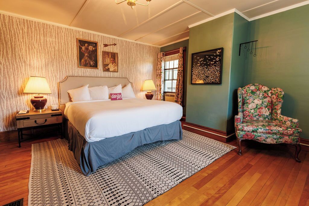 Une chambre à coucher avec un grand lit, deux tables de chevet avec des lampes, un fauteuil à motifs floraux et un tapis à dessins.