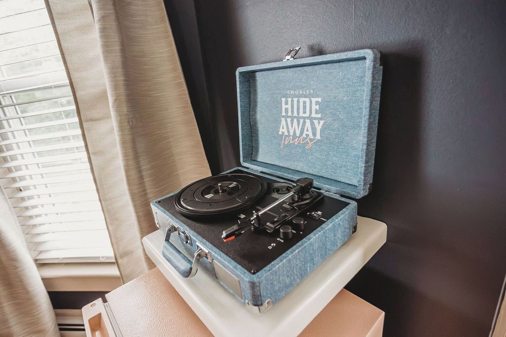 Tocadiscos antiguo de estilo vintage en estuche azul con la inscripción 'CROSLEY HIDE AWAY Inns' en la tapa, colocado sobre una superficie blanca cerca de una ventana con persianas.