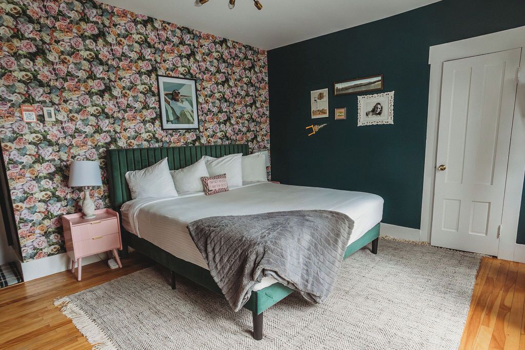 A bedroom with a floral wallpaper, a bed with a green headboard, a bedside table with a lamp, and a door.