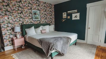 A bedroom with a floral wallpaper, a bed with a green headboard, a bedside table with a lamp, and a door.