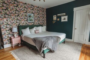 Un dormitorio con papel tapiz floral, una cama con cabecera verde, una mesita de noche con lámpara y una puerta.