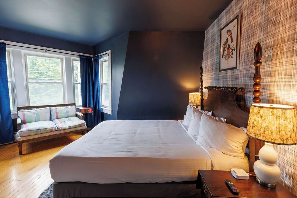 Een hotelkamer met een groot bed, een nachtkastje met lamp, een bank met kussens, en een raam met gordijnen.
