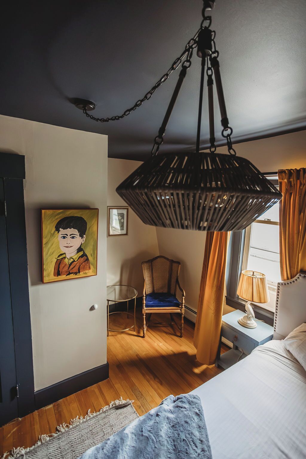 Een slaapkamer met een bed, een hanglamp, een stoel, een schilderij en een raam met gordijnen.
