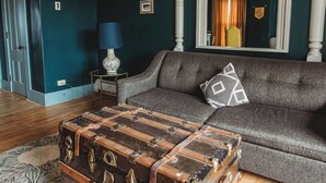 A living room with a vintage trunk as a coffee table, a grey sofa, and a mirror reflecting a room with a bicycle. - Hideaway Inn Franconia Notch (Sugar Hill)
