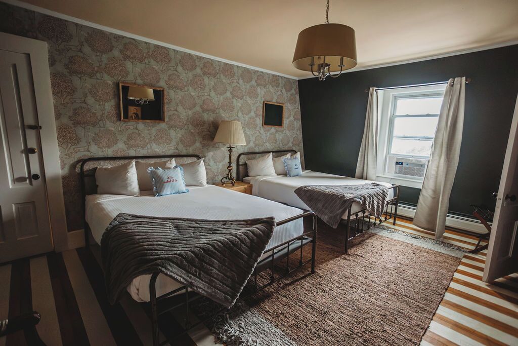 A bedroom with two beds, a chandelier, a window with curtains, and a patterned wallpaper.