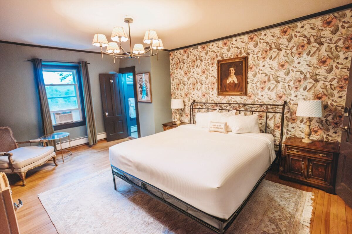 A bedroom with a large bed, a chandelier, a floral wallpaper, and a view through a window.