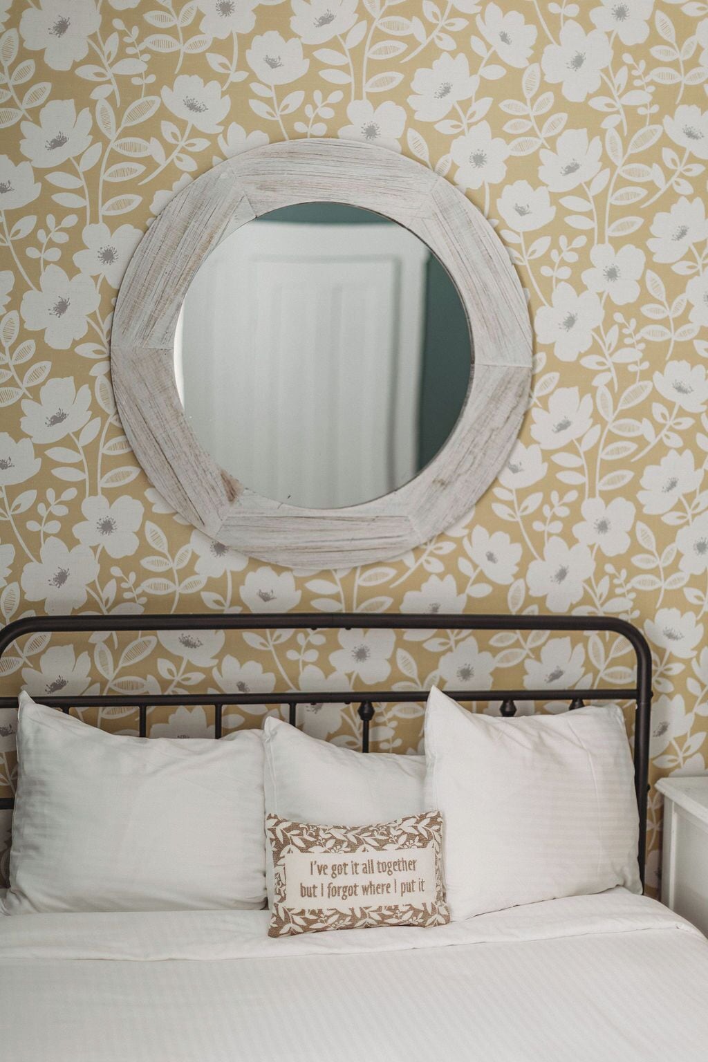 Uma cama com roupa de cama branca, um espelho redondo na parede e um papel de parede com padrão floral.