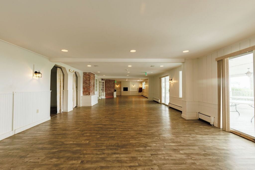 A long, open hallway with wooden flooring, white walls, and large windows on one side.