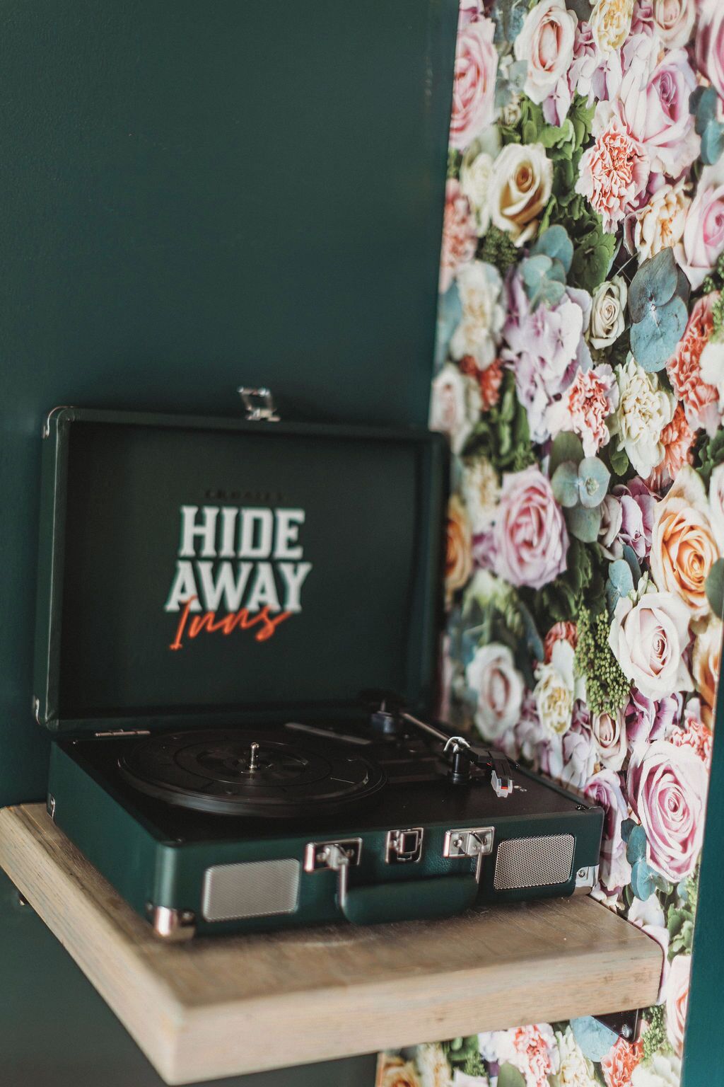 Un giradischi con il marchio 'HIDE AWAY Inns' sul coperchio, posizionato su una mensola di legno con uno sfondo a carta da parati floreale.
