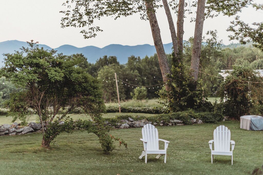 Uma área externa gramada com duas cadeiras Adirondack brancas, um muro de pedra e uma guirlanda de luzes.