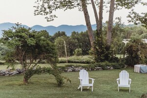 A grassy outdoor area with two white Adirondack chairs, a stone wall, and a string of lights.