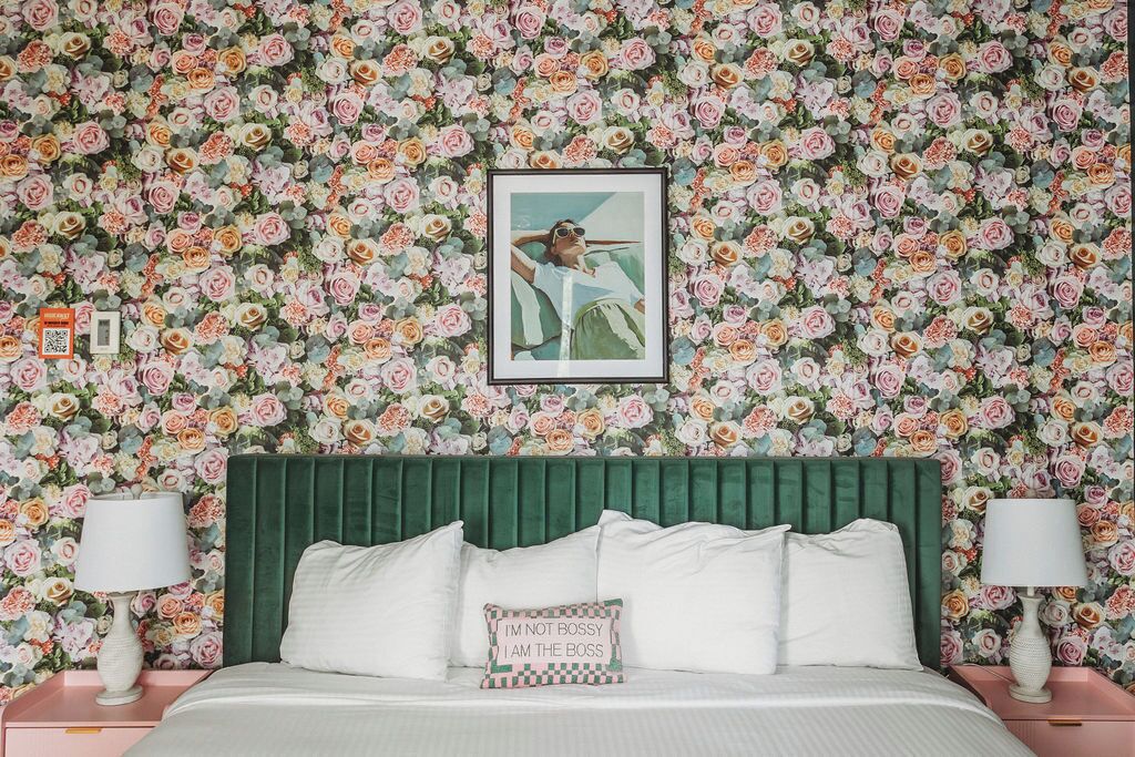 A bedroom with a floral wallpaper, a green upholstered headboard, white bedding, and a framed picture on the wall.