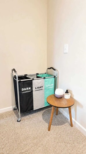 A corner of a room with a laundry cart labeled 'DARK LAUNDRY' and 'COLOR LAUNDRY,' a small round table with a white speaker and a container, and a beige carpet. - Modern Greenville Townhome 3BR, 3BA, Arden Woods (Greenville)