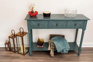 Een blauwgroene console tafel met drie lades, een plank en een rieten stoel met een blauwe deken.