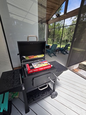 A barbecue grill with a toolset on a wooden deck with a view of a lake and chairs.