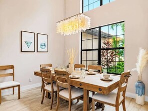 A dining area with a wooden table and chairs, a chandelier, and a large window with a view of greenery. - THE Luxury Villa, Gulf Access, Pool, Spa, Tiki Hut (Cape Coral)
