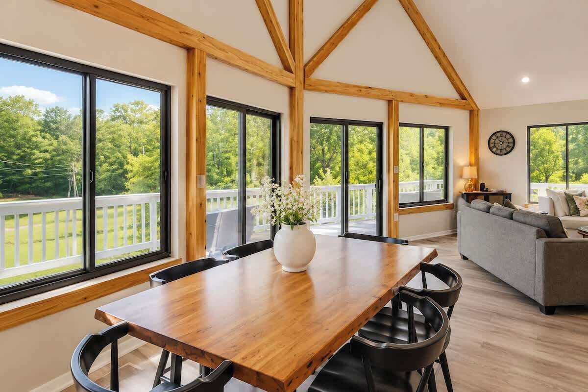 A spacious room with a wooden dining table, black chairs, and a sofa. Large windows offer a view of a green landscape.