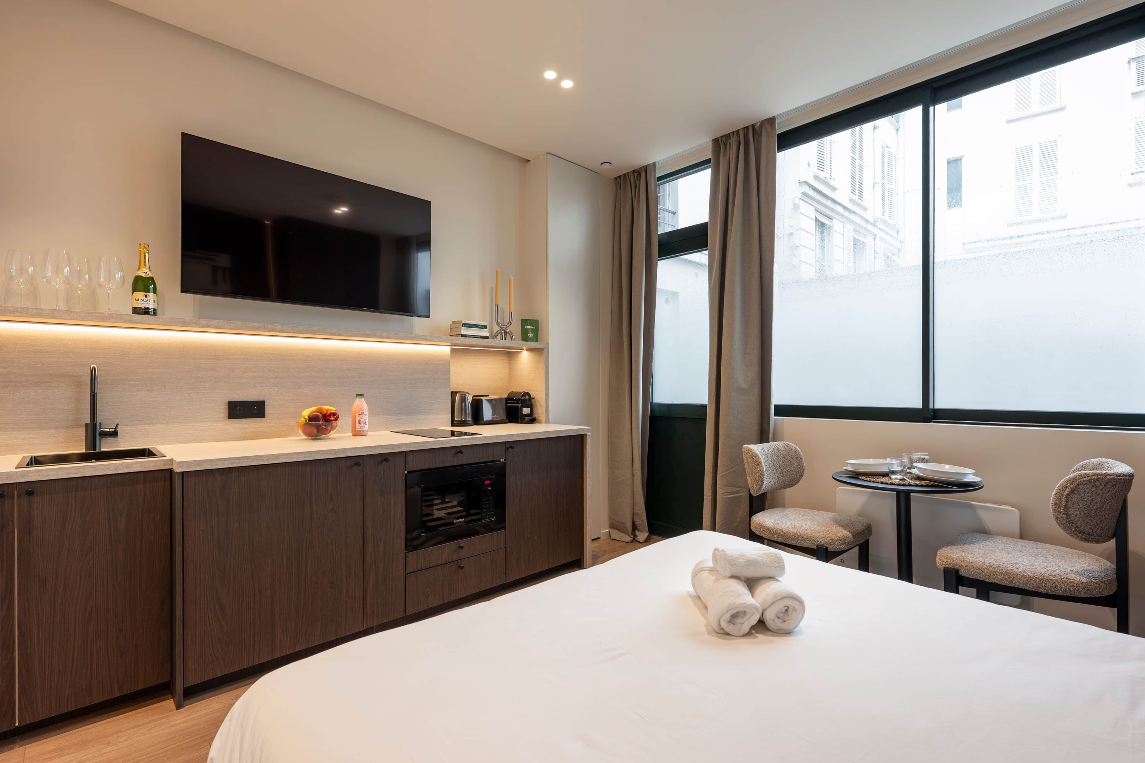Habitación de hotel moderna con kitchenette, cama con toallas, televisor y vista a la ciudad.