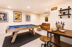 A pool table with a blue felt top, a sofa with a colorful throw blanket, a bar area with a neon sign, and a wall-mounted shelf with bottles. - Cape Coral Luxury 5BR Pool, Spa, Dock, Game Room (Cape Coral)
