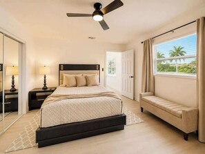 Une chambre à coucher avec un lit, des tables de chevet, un ventilateur de plafond, une fenêtre avec des rideaux et une vue sur des palmiers.