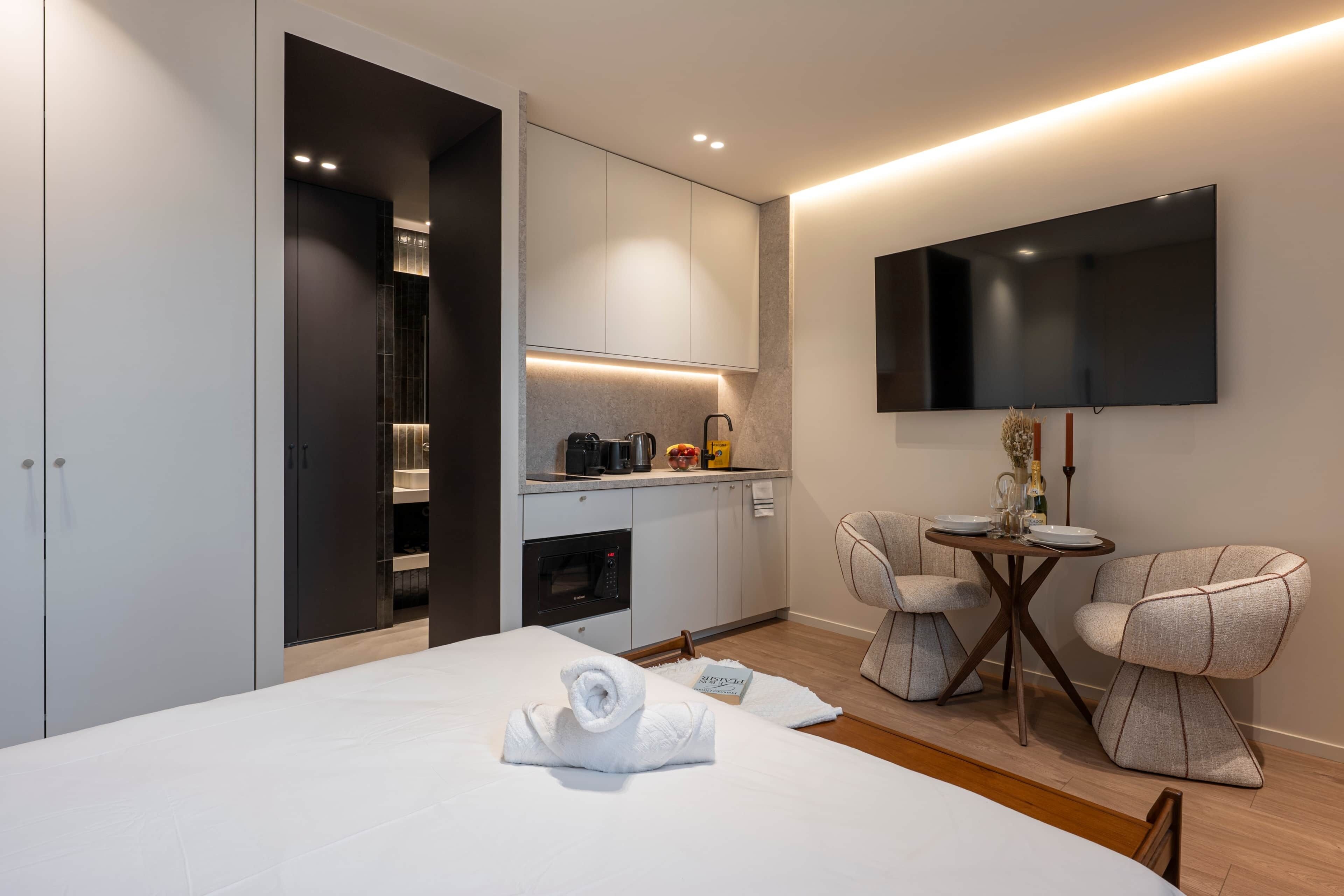A modern hotel room with a bed, a small kitchenette, a flat-screen TV, and a seating area with a table and chairs.