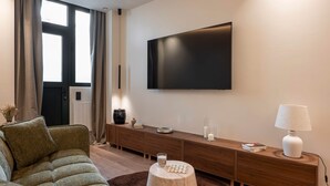 A modern living room with a large flat-screen TV mounted on the wall, a brown sofa, a wooden console table with decorative items, and a round wooden stool. - Deluxe Suite between Concorde and Vendôme (Paris)
