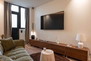 A modern living room with a large flat-screen TV mounted on the wall, a brown sofa, a wooden console table with decorative items, and a round wooden stool. - Deluxe Suite between Concorde and Vendôme (Paris)