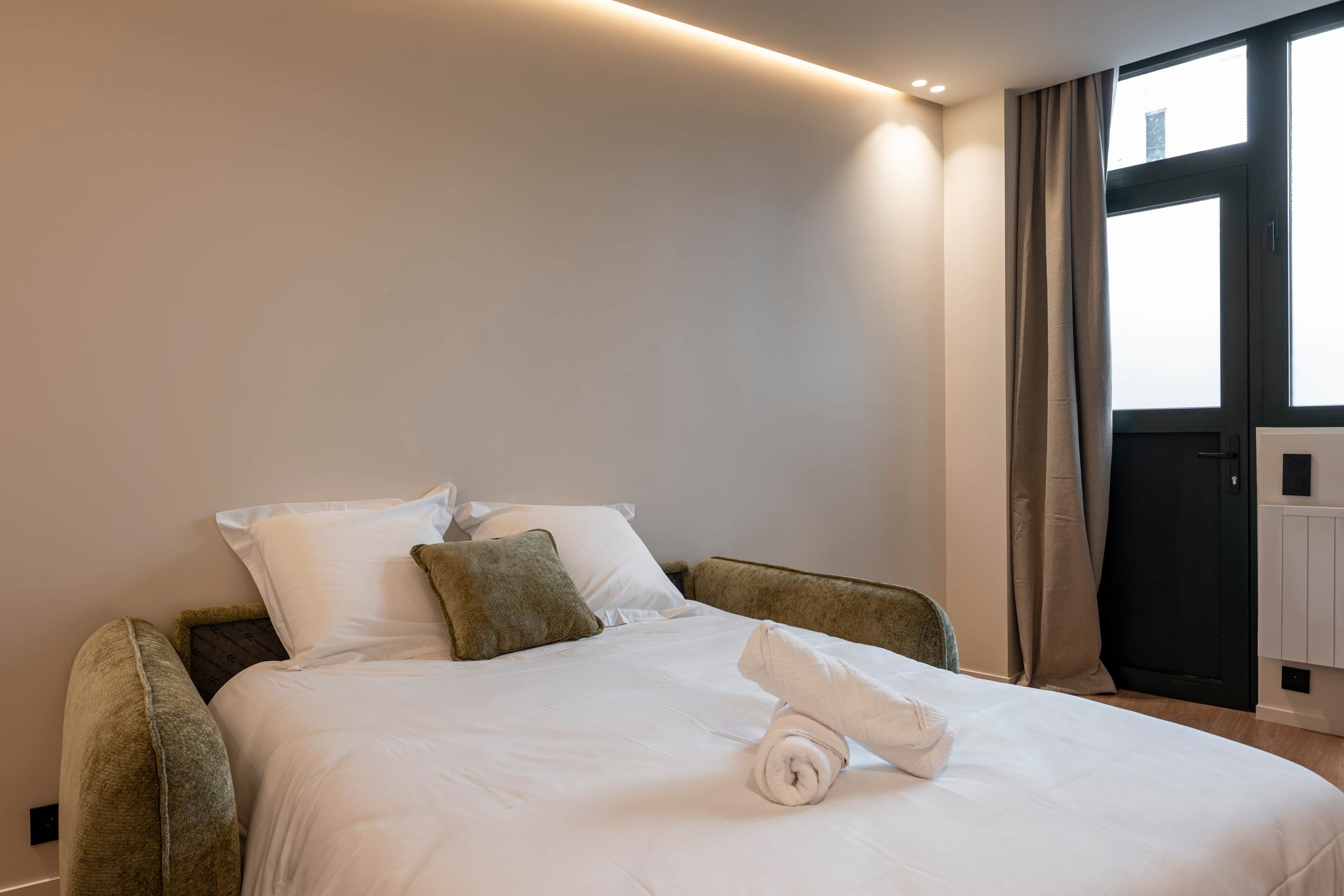 A hotel room with a bed, pillows, a rolled towel, and a window with curtains.