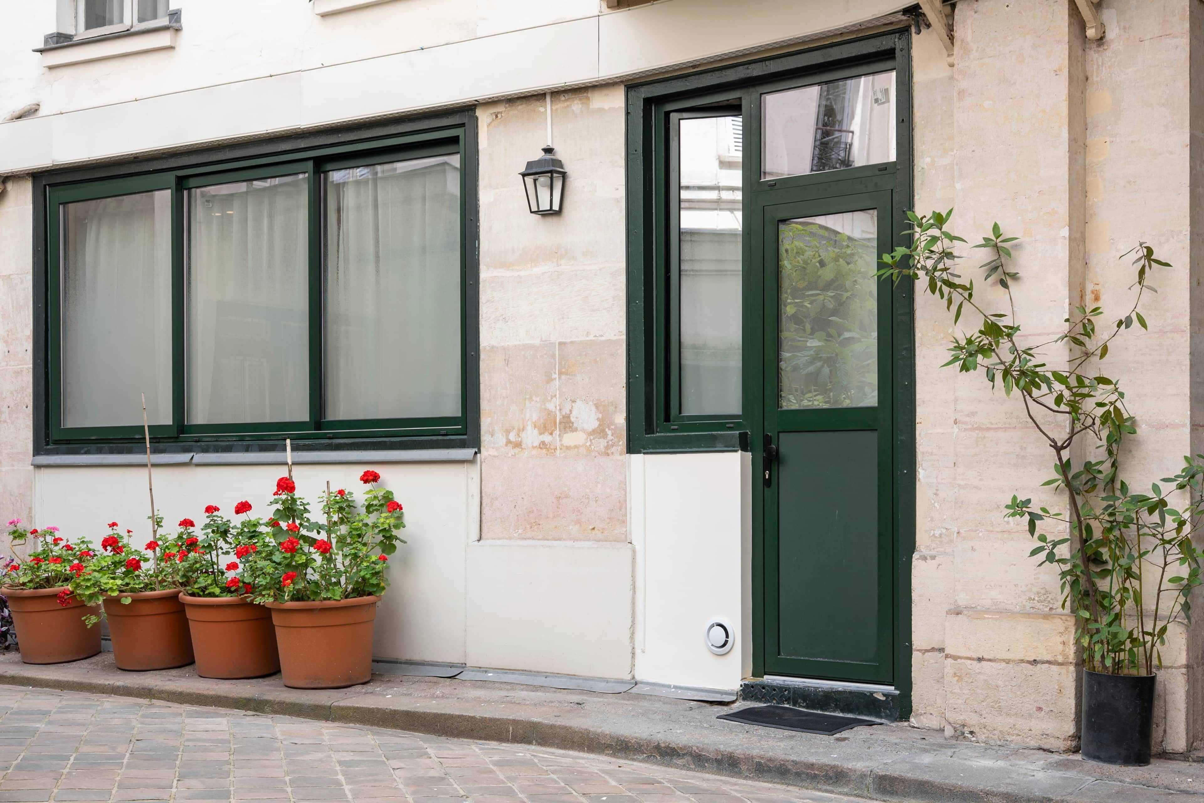 A building with a green door, a window with curtains, and potted red flowers on the ground.