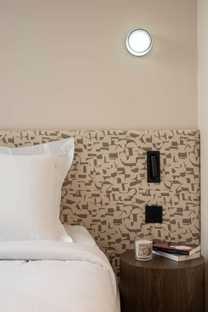 Ein ordentlich bezogenes Bett mit weißen Leinen, eine Wand mit Muster, ein kleiner Nachttisch mit Kerze und Buch.