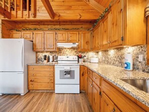 A kitchen with wooden cabinets, a white refrigerator, a stove, and a granite countertop. - Mountaintop Log Cabin with Hot Tub, Pool & Fire Pit (Sevierville)