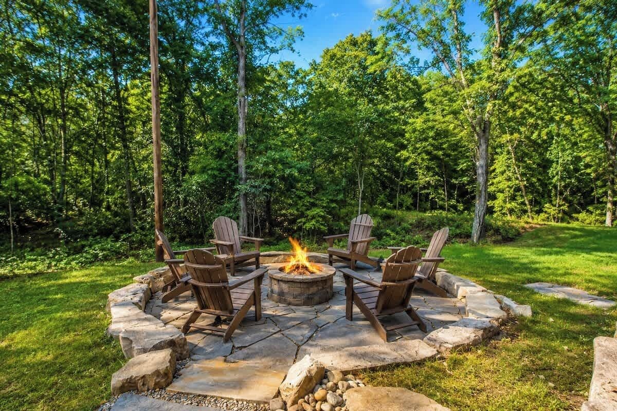 A circular stone fire pit with four Adirondack chairs around it, set in a grassy area with trees in the background.