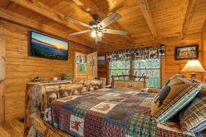 A log cabin bedroom with a bed, a ceiling fan, a window with curtains, and a painting on the wall. - Mountaintop Log Cabin with Hot Tub, Pool & Fire Pit (Sevierville)