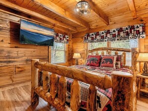 Ein Holzhütten-Interieur mit einem Bett, Holzwänden, einem montierten Fernseher und einem Fenster mit Jalousien.