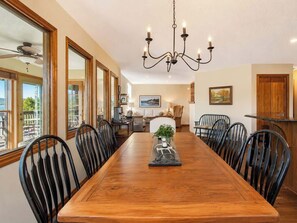 A dining room with a wooden table, black chairs, a chandelier, and a view of the outdoors through large windows.