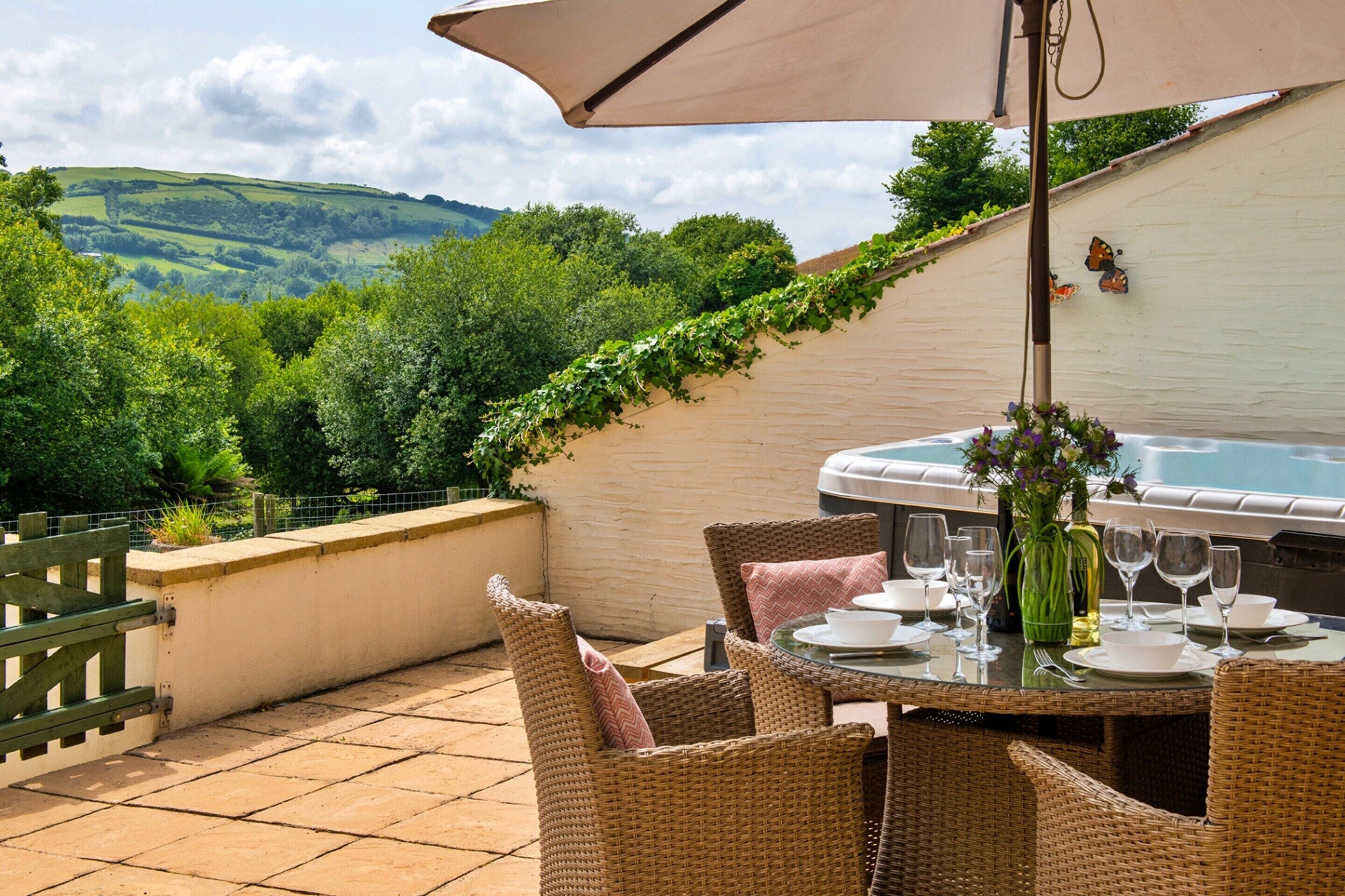 An outdoor patio with wicker furniture, a table set for dining, a hot tub, and a view of green hills.