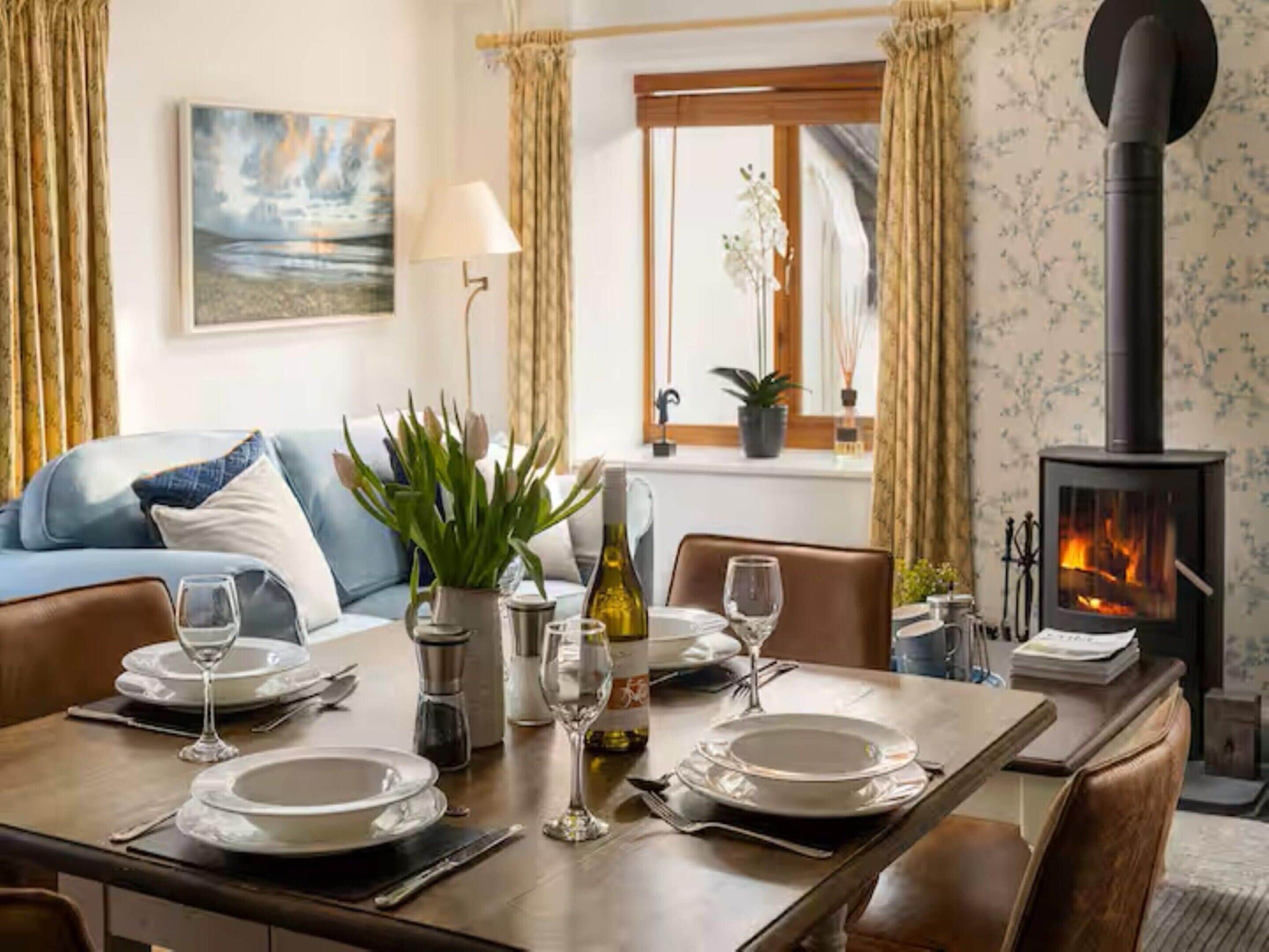 A dining area with a table set for two, a sofa, a painting, a window with curtains, and a wood-burning stove.