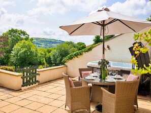 An outdoor patio with a table set for dining, surrounded by wicker chairs, a large umbrella, and a hot tub. - Coulscott Stay for Four (Ilfracombe)