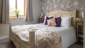 Ein Schlafzimmer mit einem Bett, Vorhängen, einem Fenster, einem Nachttisch und einer Wand mit floralem Tapetenmuster.