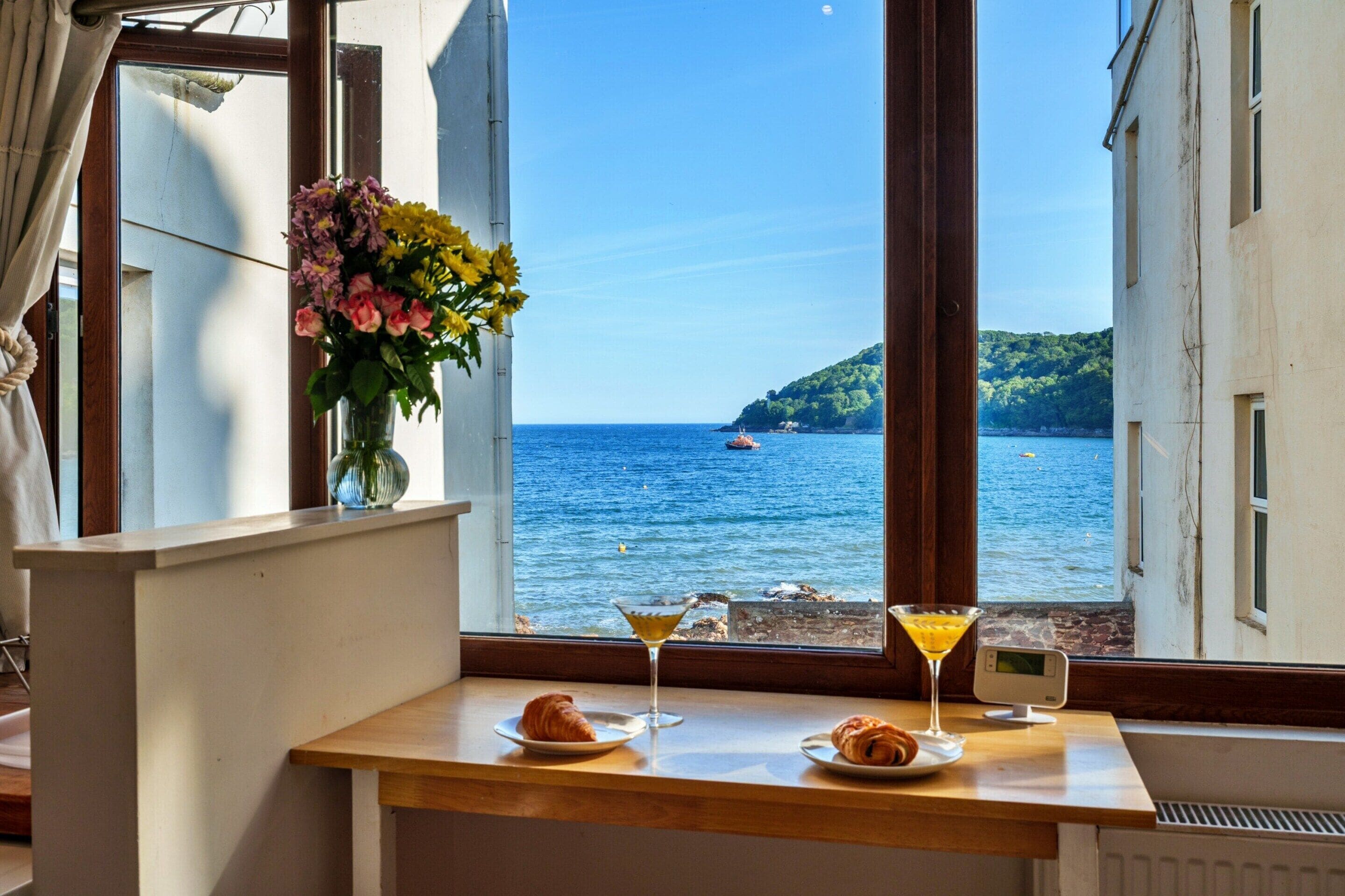 A table with croissants and a martini glass, a vase with flowers, and a view of the sea through a window.