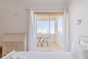 A hotel room with a bed, a wooden dresser, a balcony with a table and chair, and a view of buildings. - #416 Spacious Villa | Private Pool, BBQ, and Parking (Albufeira)