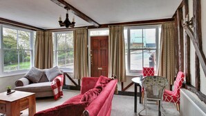 Una sala de estar acogedora con un sofá, un sillón y una mesita. Hay cortinas en las ventanas y se ve una puerta.